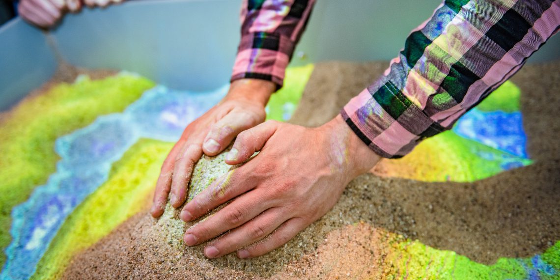 Aus Sand kann im Labor ein Höhenprofil aufgeschichtet werden. Ein Scanner erfasst es und projiziert farbcodierte Höhenlinien darauf. Mit Handbewegungen kann virtueller Regen ausgelöst werden. Bild: Damian Gorczany